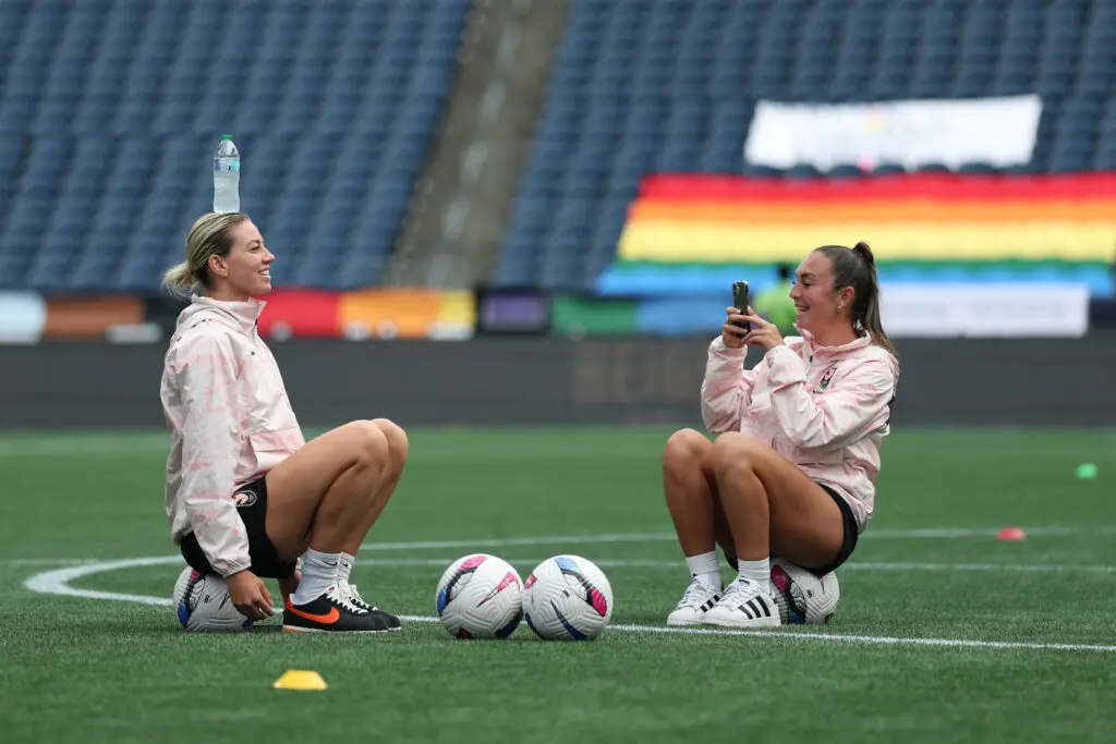 Angel City defender Alanna Kennedy balances a water bottle on her head while midfielder Katie Zelem snaps a photo before a 2025 NWSL match.