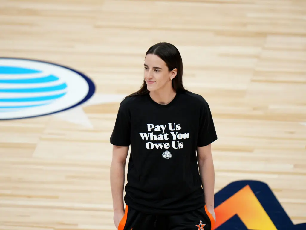 Indiana Fever guard Caitlin Clark stands on the 2025 WNBA All-Star court wearing a T-shirt that reads "Pay Us What You Owe Us."