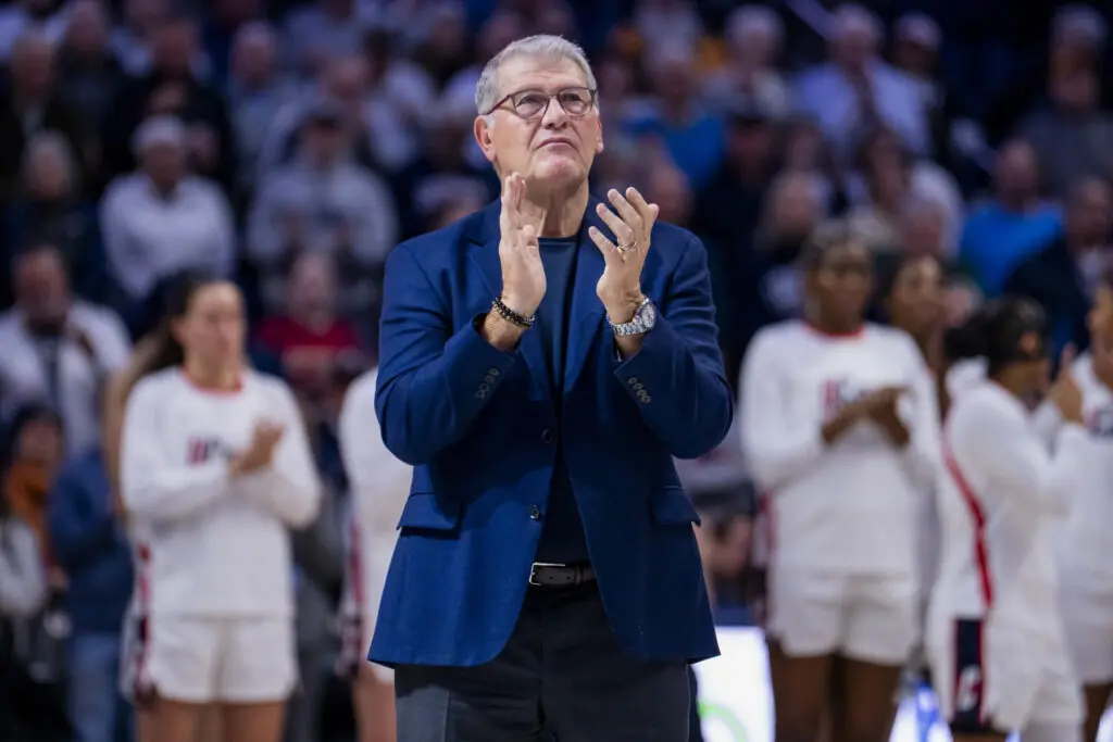 UConn head coach Geno Auriemma applauds a tribute video for past teams before a 2025/26 NCAA basketball game.