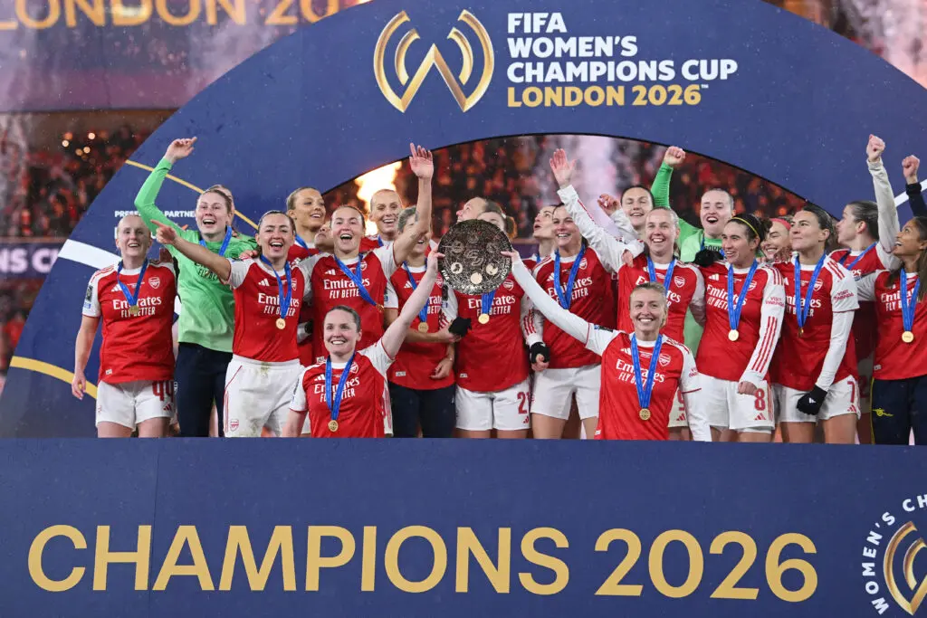 Arsenal captain Kim Little and vice-captain Leah Williamson lift the 2026 FIFA W Champions Cup while the team cheers on the podium.