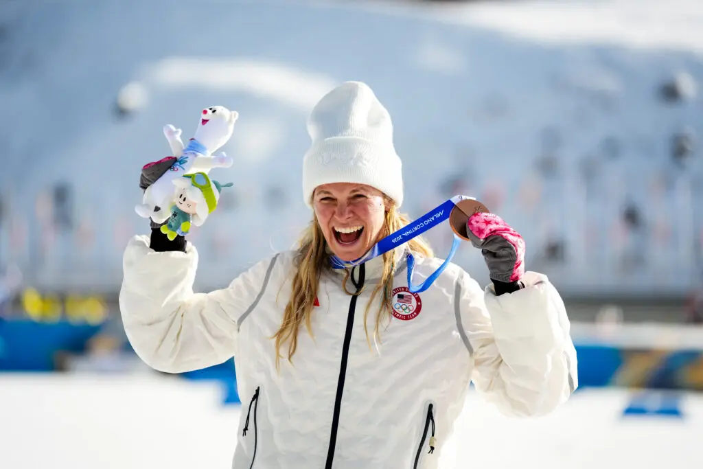 USA cross-country ski star Jessie Diggins grins while holding up her 10km bronze medal during the medal ceremony at the 2026 Winter Olympics.