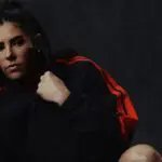 Kelsey Plum poses in front of a dark backdrop in an Adidas track top.