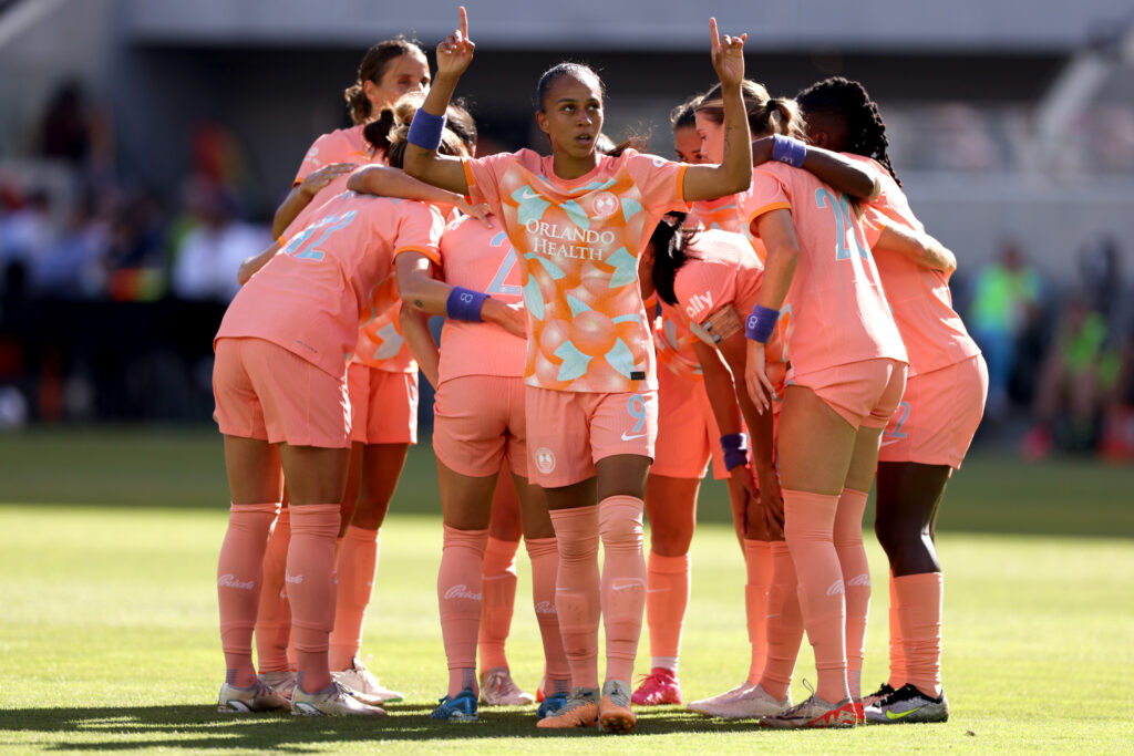 Adriana points up as her Orlando Pride celebrates a goal with a huddle.