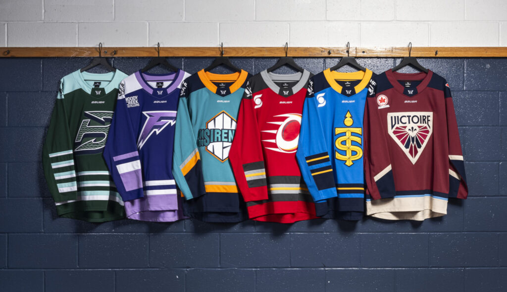 PWHL hockey jerseys hanging against a blue and grey brick wall.