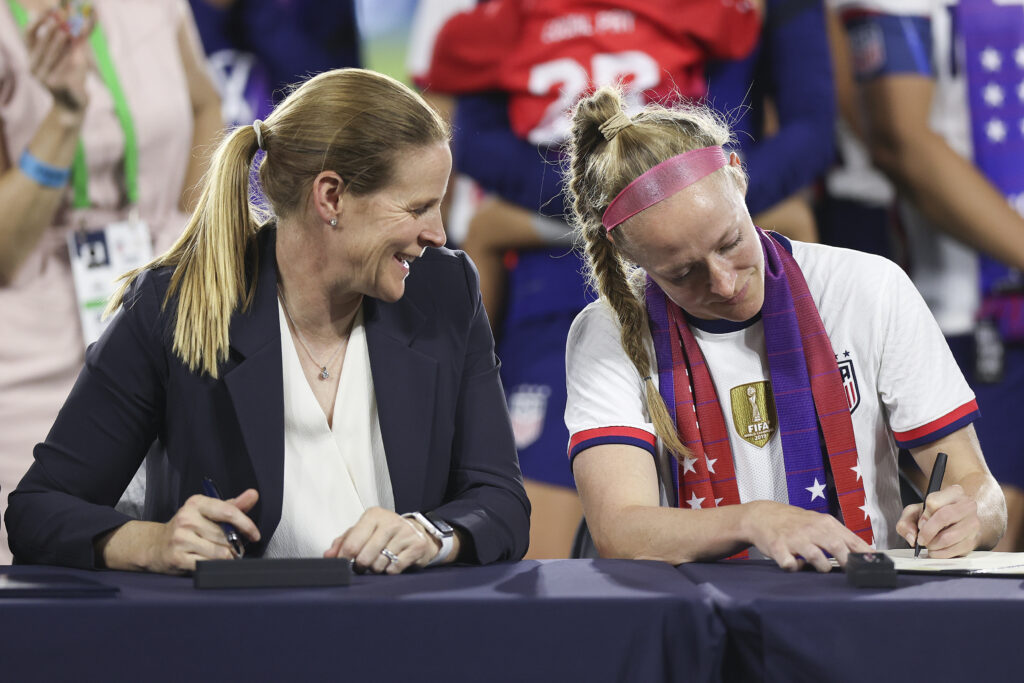 USWNT Players Association president Becky Sauerbrunn signs the 2022 equal pay CBA as US Soccer president Cindy Parlow Cone looks on.