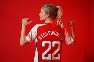 Former USWNT and NWSL midfielder Jenna Nighswonger points to the Arsenal crest on her jersey while posing against a red backdrop.