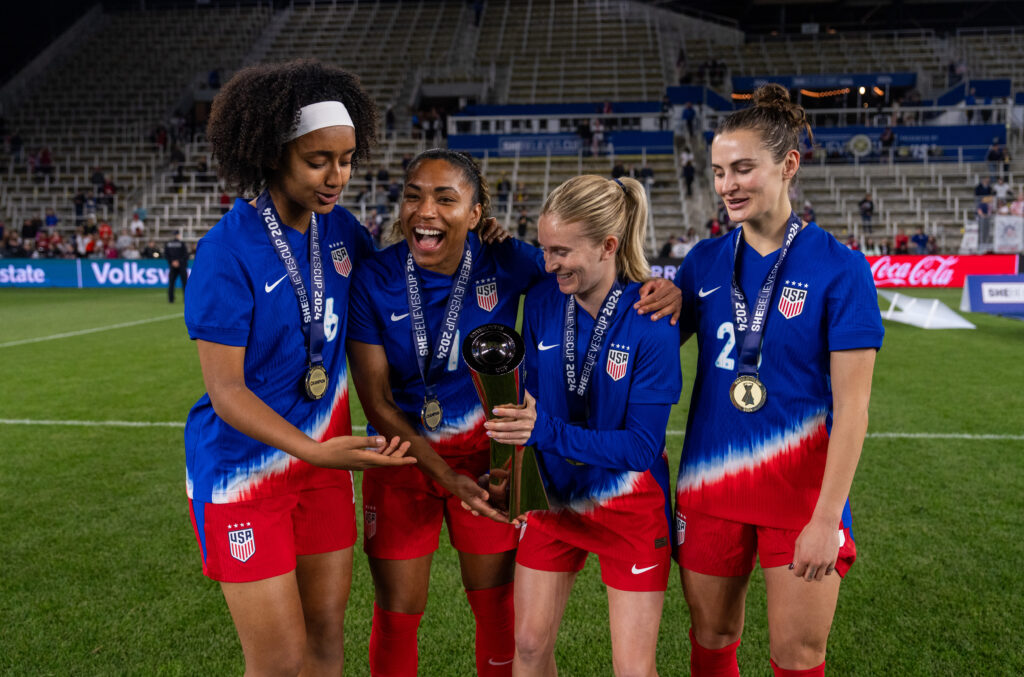 USWNT players Lily Yohannes, Cat Macario, Jenna Nighswonger, and Emily Fox hold the 2024 SheBelieves Cup trophy.