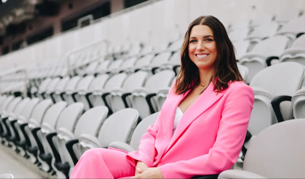 San Diego Wave minority owner Alex Morgan wears a pink suit and smiles while sitting in gray stadium seats.