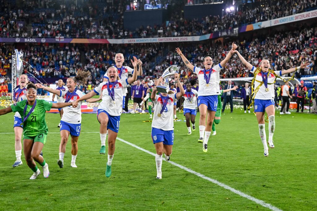 England players run with the 2025 Euro trophy to celebrate their title win with the Lionesses' fan section.