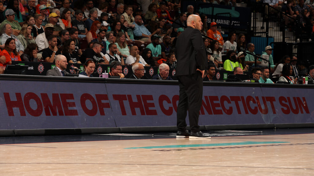 Head Coach Rachid Meziane stands in front of a sideline sign reading "Home of the Connecticut Sun" during a 2025 WNBA game.