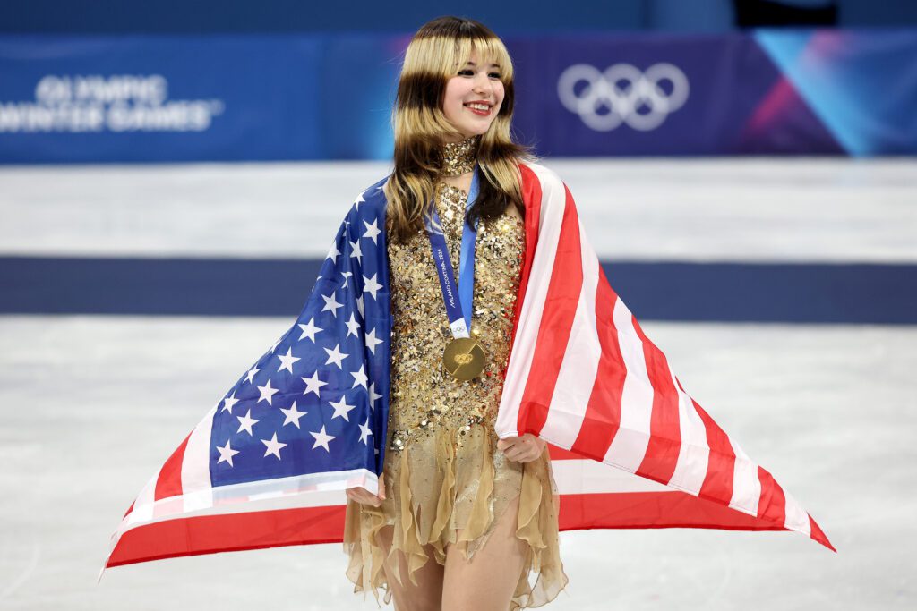 USA figure skater Alysa Liu smiles wearing her gold medal with a US flag draped over her shoulders on the 2026 Olympic podium.