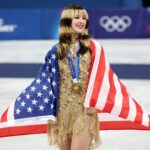 USA figure skater Alysa Liu smiles wearing her gold medal with a US flag draped over her shoulders on the 2026 Olympic podium.