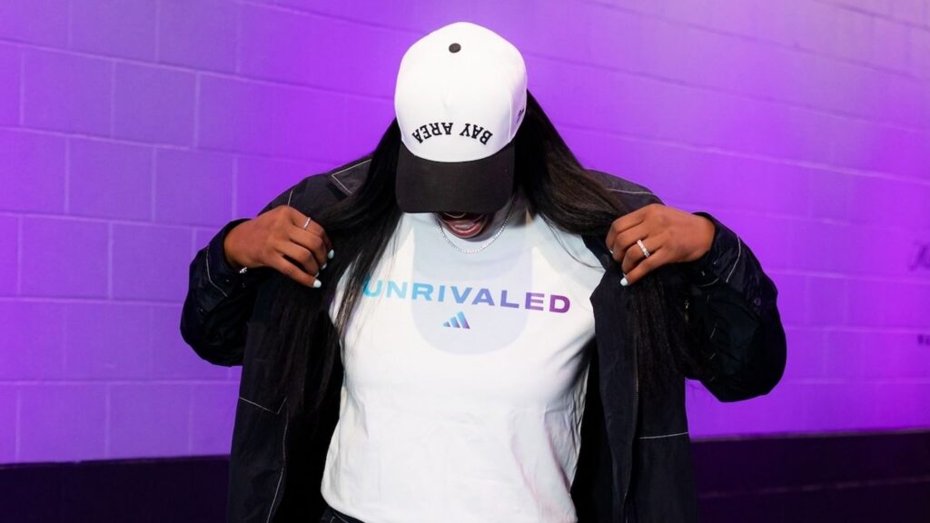 Model in a baseball cap and black jacket showing off an Unrivaled x Adidas shirt against a purple brick background.